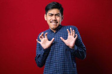 Genç yakışıklı Asyalı bir adam kırmızı arka plan üzerinde gündelik gömleğini giyiyor. Avuçlarını hareket ettiriyor. Korkmuş ve iğrenç bir ifadeyle reddediyor. Dur ve yasakla.