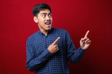 Genç yakışıklı Asyalı adam kırmızı arka planda rahat gömlekler giyiyor. Yüzünde kocaman bir gülümseme var. Parmağıyla kameraya bakıyor..