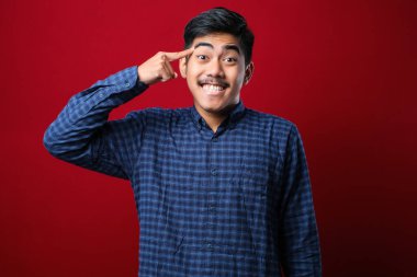 Kırmızı arka planda rahat gömlekler giyen genç yakışıklı bir adam. Bir parmağıyla gülümseyerek, harika bir fikir ya da düşünceyle, iyi bir hafızayla.