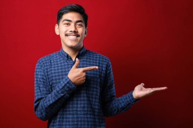 Genç, yakışıklı bir adam, sıradan gömlekler giyiyor, avuç içini gösteriyor ve baş parmaklarıyla iyi bir jest yapıyor, kırmızı arka planda mutlu ve neşeli gülümsüyor.
