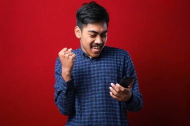 Akıllı telefon kullanan genç Hintli adam izole edilmiş kırmızı arka planda gururla bağırıyor ve zaferi ve başarıyı kutluyor çok heyecanlı, coşkulu bir duygu.