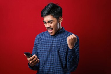 Akıllı telefon kullanan genç Hintli adam izole edilmiş kırmızı arka planda gururla bağırıyor ve zaferi ve başarıyı kutluyor çok heyecanlı, coşkulu bir duygu.