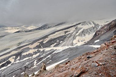 Dağ yamaçları ve karla kaplı siyah volkanik kayalar. Düşük bulutlar azgın tepelerin üzerinde