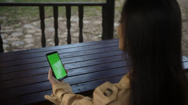 Young woman at nature sitting on a bench using with Green Mock-up Screen Smartphone in Horizontal Landscape Mode. Girl Using Mobile Phone, Browsing Internet, Watching Content, Videos or Blogs. POV plan. Technology and life style concept