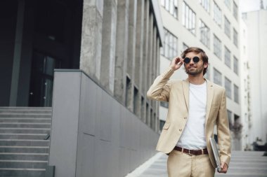 Siyah yuvarlak güneş gözlüğünü ayarlayan ve elinde kablosuz laptop tutan başarılı iş adamının ön görüntüsü. Modern teknolojiyi iş için kullanan şık bej takım elbiseli yakışıklı bir erkek..