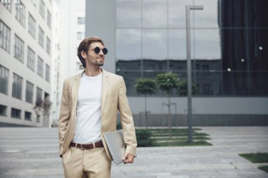 Modern ofis binasının önünde dizüstü bilgisayarı olan çekici sakallı adamın portresi. Genç işadamı takım elbise ve güneş gözlüğü takıyor..