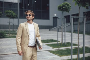 Moda takım elbiseli ve güneş gözlüklü, modern dizüstü bilgisayarını ofis dışında kullanan kendine yeten genç bir adam. Sakallı iş adamı dışarıda birini bekliyor..