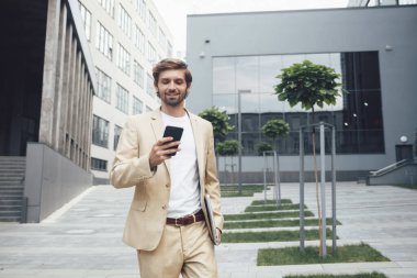 Resmi takım elbiseli kendine güvenen genç işadamı, elinde bilgisayarla ofis binasının yanında dururken modern akıllı telefon kullanıyor. İnsanlar ve teknoloji kavramı.