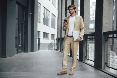 Bir elinde modern akıllı telefon, diğer elinde taşınabilir dizüstü bilgisayar olan şık takım elbiseli yakışıklı bir adam. Ofisin yakınında duran sakallı iş adamı. Uzak çalışma.