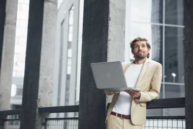 Moda takım elbiseli sakallı adam dışarıda dizüstü bilgisayarla video konferansı yapıyor. Genç işadamı, modern kulaklıklarla meslektaşlarını dinlerken yukarıdan bakıyor..