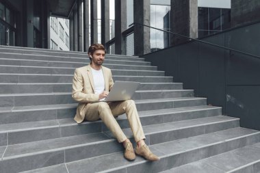 Resmi giysiler içinde çekici, mutlu bir adam merdivenlerde oturuyor ve kişisel dizüstü bilgisayarla çalışıyor. Uzaklık çalışması.