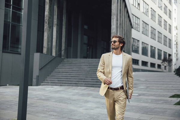 Счастливый бородатый мужчина ходит по улице с портативным ноутбуком в руке. Уверенный мужчина в деловом костюме и стильных солнечных очках. Внешний вид бизнес-центра.
