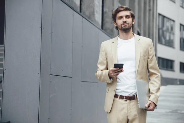Thoughtful bearded man dressed in trendy beige suit holding smartphone and wireless laptop, and looking aside. Busy businessman thinking about work out of the office.