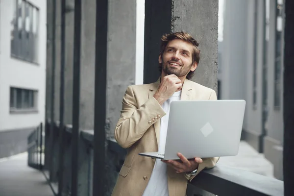 Thoughtful bearded man in formal outfit standing near handrail with opened laptop outdoors. Young businessman looking for new ideas in working project.