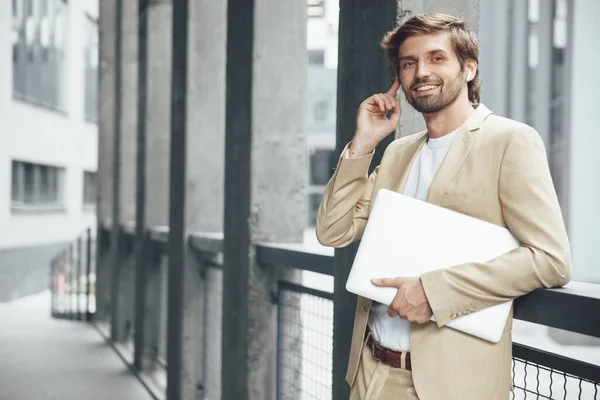 Smiling young man with stylish haircut and beard touching with finger one earphones and looking at camera. Bearded businessman leaning on handrail and holding personal laptop.