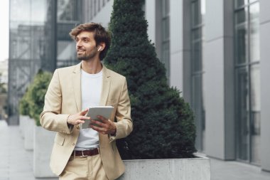 Elinde dijital tabletle caddede duran şık takım elbiseli yakışıklı adamın portresi. Genç iş adamı kablosuz kulaklık kullanıyor. Başarılı kariyer kavramı.