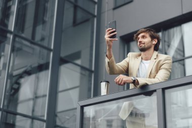 Resmi giyinmiş yakışıklı bir adamın ofis merkezinin balkonunda dikilirken akıllı telefondan görüntülü konuşma yapması. Sakallı erkek, uzak iş için modern aletler kullanıyor..