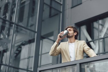 Kendine güvenen, şık takım elbiseli genç işadamı termo bardaktan sıcak kahve içerken dışarıda dikiliyor. Mutlu sakallı adam ofisten çıkıp temiz hava alıyor..
