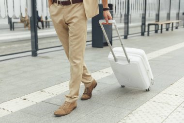 Moda kıyafetli genç adam otobüs durağında beyaz bavuluyla ayakta duruyor. Erkek turist dışarıda toplu taşıma bekliyor. Kapat..