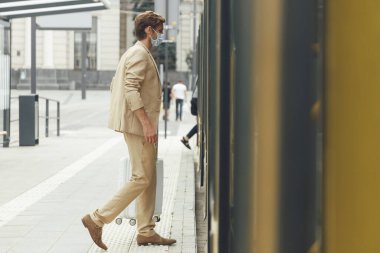 Tıbbi maskeyle tramvaya binen yakışıklı adamın yan görüntüsü. Genç sakallı adam takım elbise giyiyor ve beyaz bavul taşıyor..