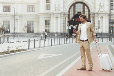 Kendine güvenen takım elbiseli bir adamın uzun bir portresi tren istasyonunun yanında duruyor ve seyyar ekrana bakıyor. İnsan kavramı, teknoloji ve yolculuk.