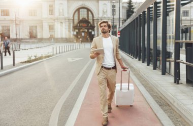 İş gezisinde dijital tablet kullanan şık takım elbiseli kendine güvenen sakallı bir adam. Ciddi bir işadamı beyaz bavulla istasyondan buraya yürüyor..