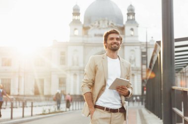 Elinde dijital tabletle dışarıda kameraya poz veren kendine yeten sakallı bir adam. İş gezisi sırasında modern aletleri kullanan genç ve mutlu bir erkek..