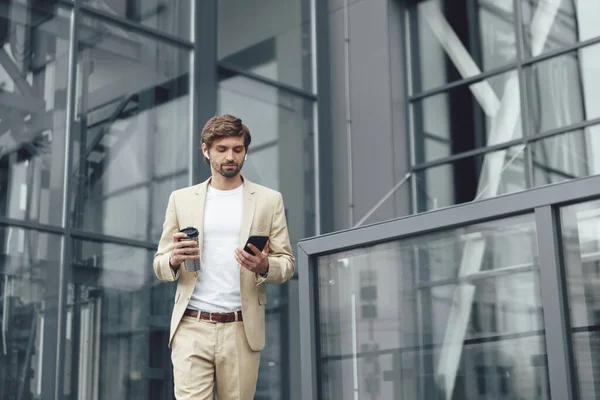 Successful businessman in suit and wireless earphones walking on street with modern smartphone and cup of coffee. Bearded man with trendy haircut using mobile outdoors.