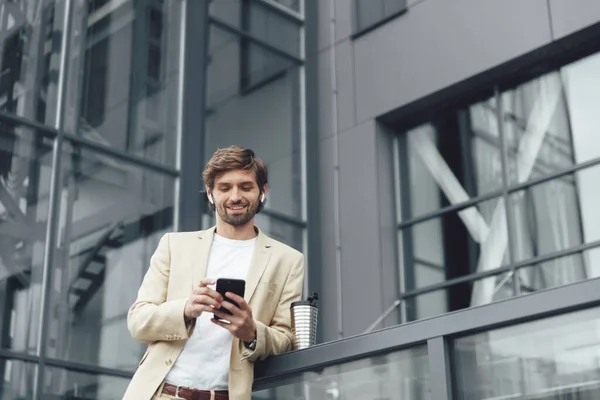 Smiling young man in formal wear leaning on railings and using modern smartphone. Thermo cup with hot coffee standing near. Business center on background.