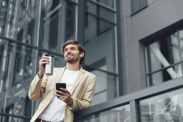 Successful man in stylish business suit holding in one hand modern smartphone and cup of coffee in another. Concept of finance, career and technology.