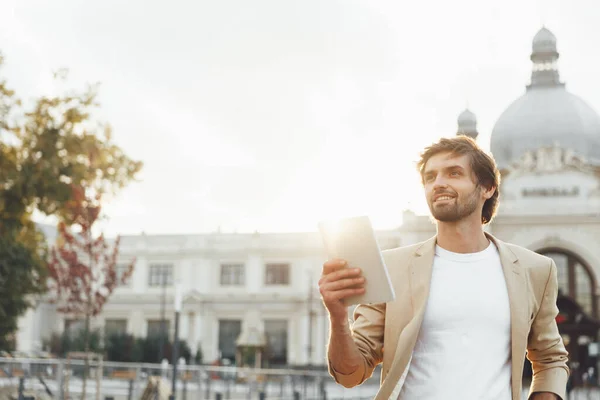 Portrait of attractive man with trendy haircut and beard using digital tablet outdoors. Businessman in suit standing near railway station and looking aside. Warm sunny day.