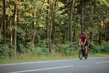 Yeşil yaz ormanlarında siyah bisiklet süren aktif giysili ve koruyucu kasklı genç sakallı bir sporcu. Temiz hava üzerinde çalışma kavramı.