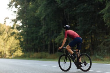 Yeşil ormanların arasında bisiklet süren kaslı, koruyucu kasklı ve aktif kıyafetli adamın arka görüntüsü. Bisiklet ve spor etkinliği kavramı.