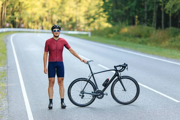 Happy muscular guy in black helmet and mirrored glasses relaxing on rad after morning ride. Cyclist in sport clothing resting with bike on asphalt road.