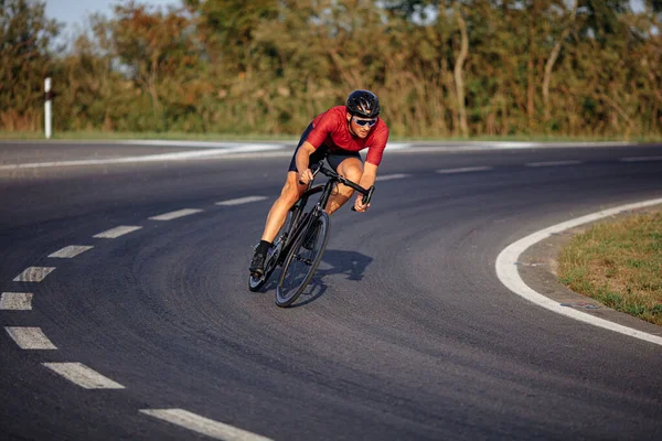 Healthy young sportsman in black helmet and protective eyewear biking on asphalt road outdoors. Muscular cyclist in action.