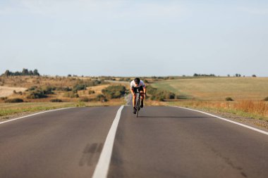 Önde, asfalt yolda bisiklet süren koruyucu kasklı ve spor elbiseli yakışıklı bir genç var. Dayanıklılık ve güç kavramı.