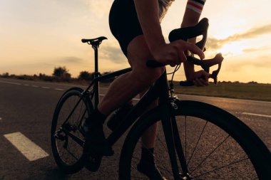 Kırsal kesimde asfalt yolda siyah bisiklet süren spor kıyafetli aktif bir adam. Arka planda inanılmaz bir gün batımı. Açık hava spor aktivitesi.