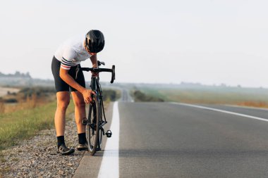 Koruyucu kasklı ve spor kıyafetli yakışıklı erkek sporcu siyah bisikletin tekerlerini kontrol ediyor. Yolda duran genç bisikletçi yarı yamuk pozisyonda..