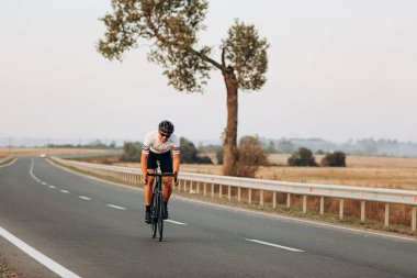 Güler yüzlü güçlü bisikletçi dışarıda siyah bisikletle spor yapmaktan zevk alıyor. Kasklı ve gözlüklü yakışıklı bir adam yaz günü asfalt yolda gidiyor..