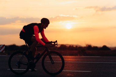 Parlak bisiklet kıyafetleri giymiş erkek sporcu akşam vakti kaldırımlı yolda yarış yapıyor. Kasklı profesyonel bisikletçi temiz havada en sevdiği hobiyi yapıyor..