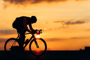 Korumalı kasklı profesyonel bisikletçinin silueti kaldırım yolundaki spor aktiviteleri için bisiklet kullanıyor. Yaz günbatımının arka planı.