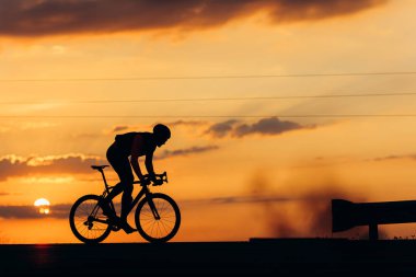 Güçlü erkek atlet silueti yolda daha yüksek hız için yarı eğimli pozisyonda bisiklet sürüyor. Günbatımında favori hobisinin keyfini çıkaran profesyonel bisikletçi..