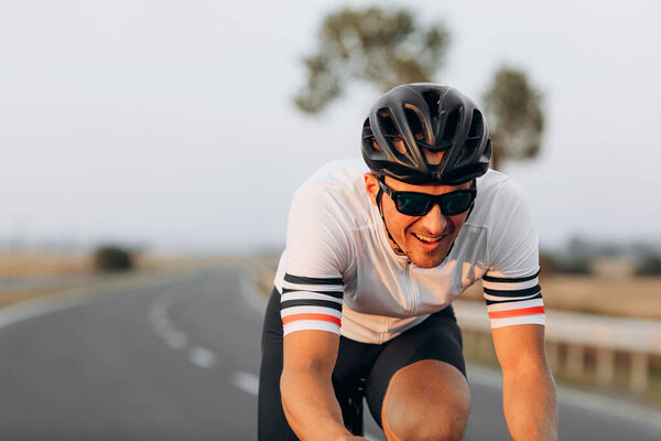 Portrait of sporty bearded man in eyewear and protective helmet riding bike outdoors. Blur background of asphalt road among fields.