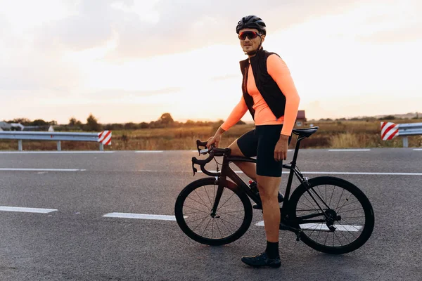 Happy young cyclist dressed in sport clothing posing with black bike on paved road. Active sportsman enjoying evening training on fresh air.