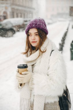 Kar yağarken sokakta duran ve elinde sıcak kahve tutan kalın giysiler giymiş çekici genç bir kadın. Kış tatili ve atmosfer kavramı.