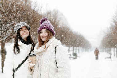 Dışarıdaki kar yağışı sırasında sıcak kahve içen sıcak kıyafetler giymiş hoş genç bayanların ön görüntüsü. Mutlu arkadaşlar sokaklarda yürüyor ve kış tatillerinin tadını çıkarıyorlar..