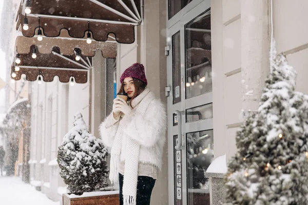 白いジャケット 赤い帽子 ニットスカーフの魅力的な若い女性は 雪の冬の日に熱い飲み物を楽しんでいます 居心地の良い街のカフェの近くでコーヒーを持っている幸せな女性 — ストック写真
