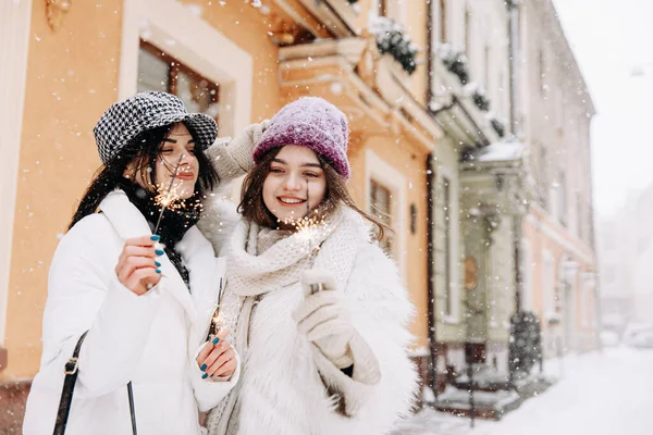 Group of two young women in winter clothing posing on camera on street with sparklers in hands. Holidays atmosphere. Friendship concept.