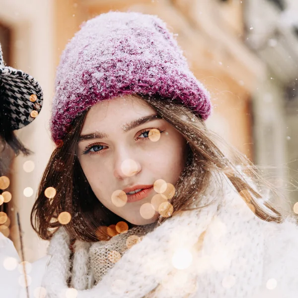 ストリートに立っている間にカメラを見てニット帽と白いジャケットで魅力的な若い女性のクローズアップ 雪の多い冬の天気 背景に輝く人 — ストック写真