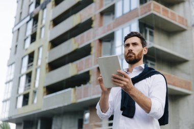 Tabletli sakallı adam bitmemiş evlerin arasında duruyor.